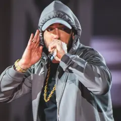 Eminem w szarej bluzie z kapturem i czapce, trzymający mikrofon i gestykulujący.