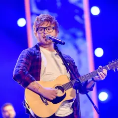 Ed Sheeran w okularach gra na gitarze akustycznej i śpiewa do mikrofonu na niebieskiej scenie.