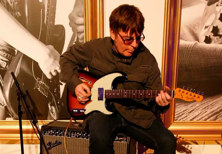 Mężczyzna w okularach gra na gitarze elektrycznej Fender, siedząc na wzmacniaczu.